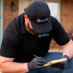 Man wearing a black Partico Solutions cap and gloves writes on a yellow notepad outdoors; brick background.