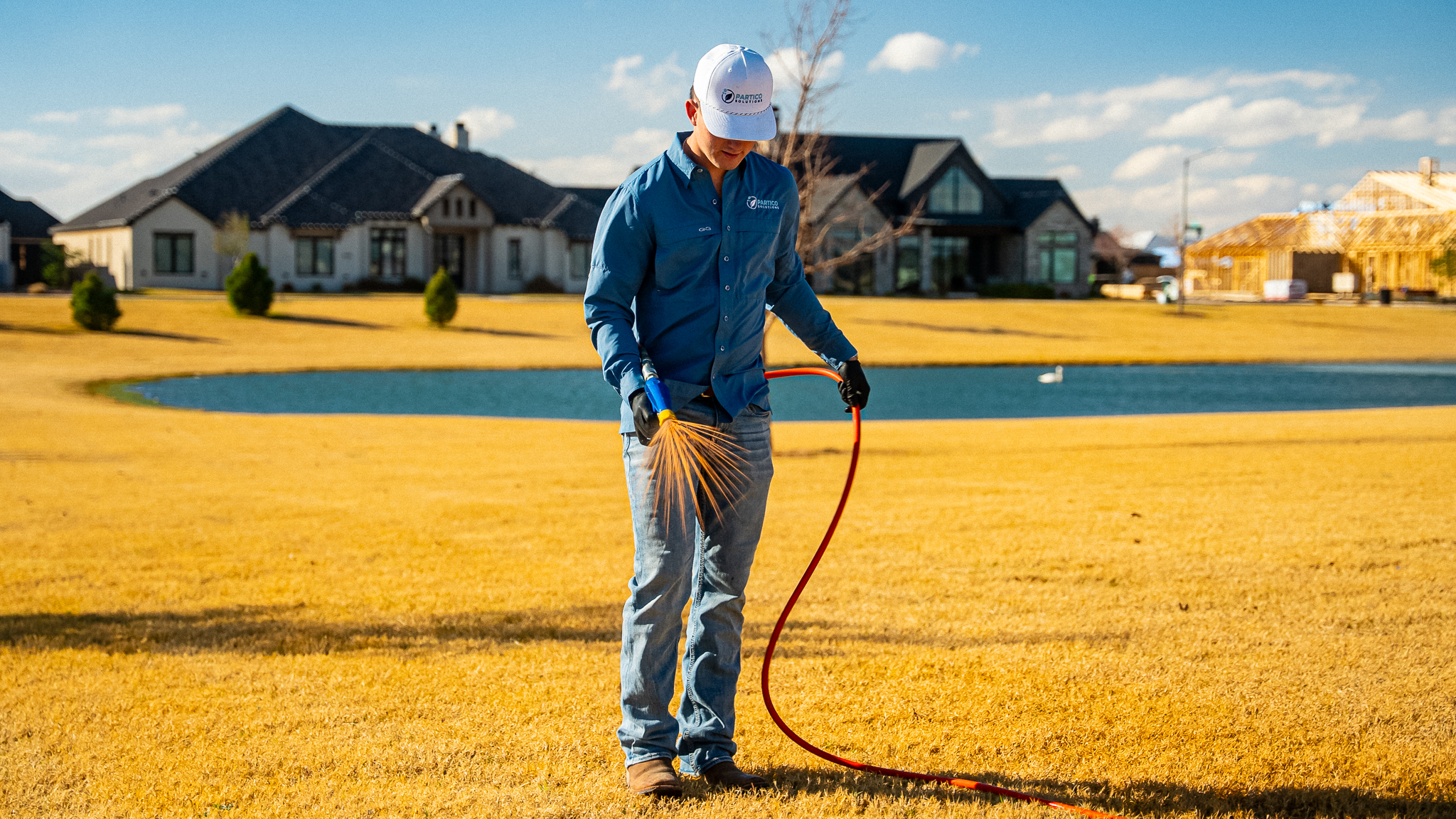 Construction worker in a blue shirt and white cap stands on a yellowed lawn near a pond, holding an orange hose and a yellow broom as houses under construction appear in the background.
