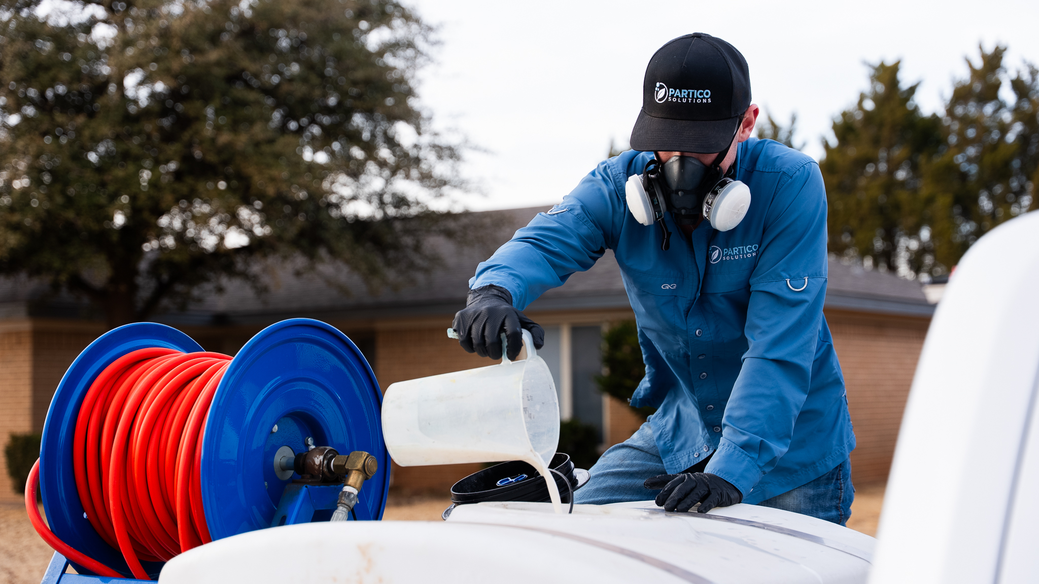 Utility worker in a blue uniform and respirator pours liquid into a white container next to a coiled red hose on a blue reel outdoors.