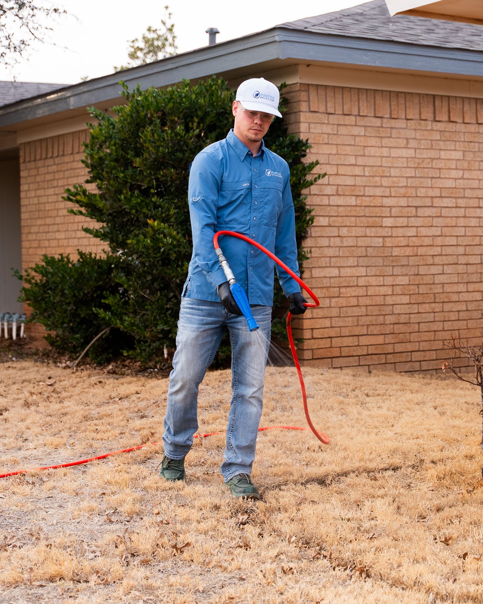 Man in a blue work shirt and white cap holds a red hose outside a brick house, spraying water on the dry lawn.