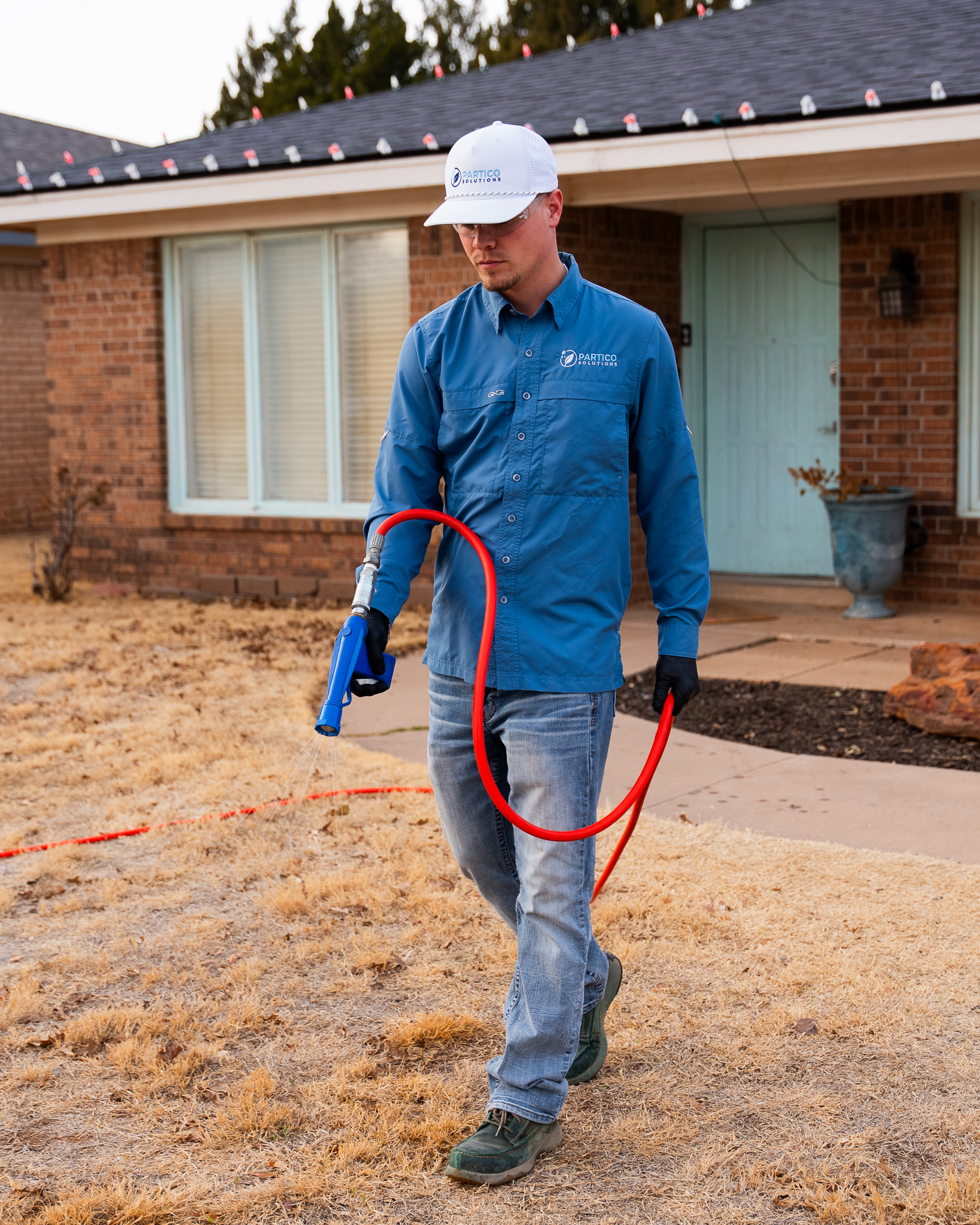 Man in a blue work shirt and white cap holding a red hose nozzle in a dry front yard of a brick house.
