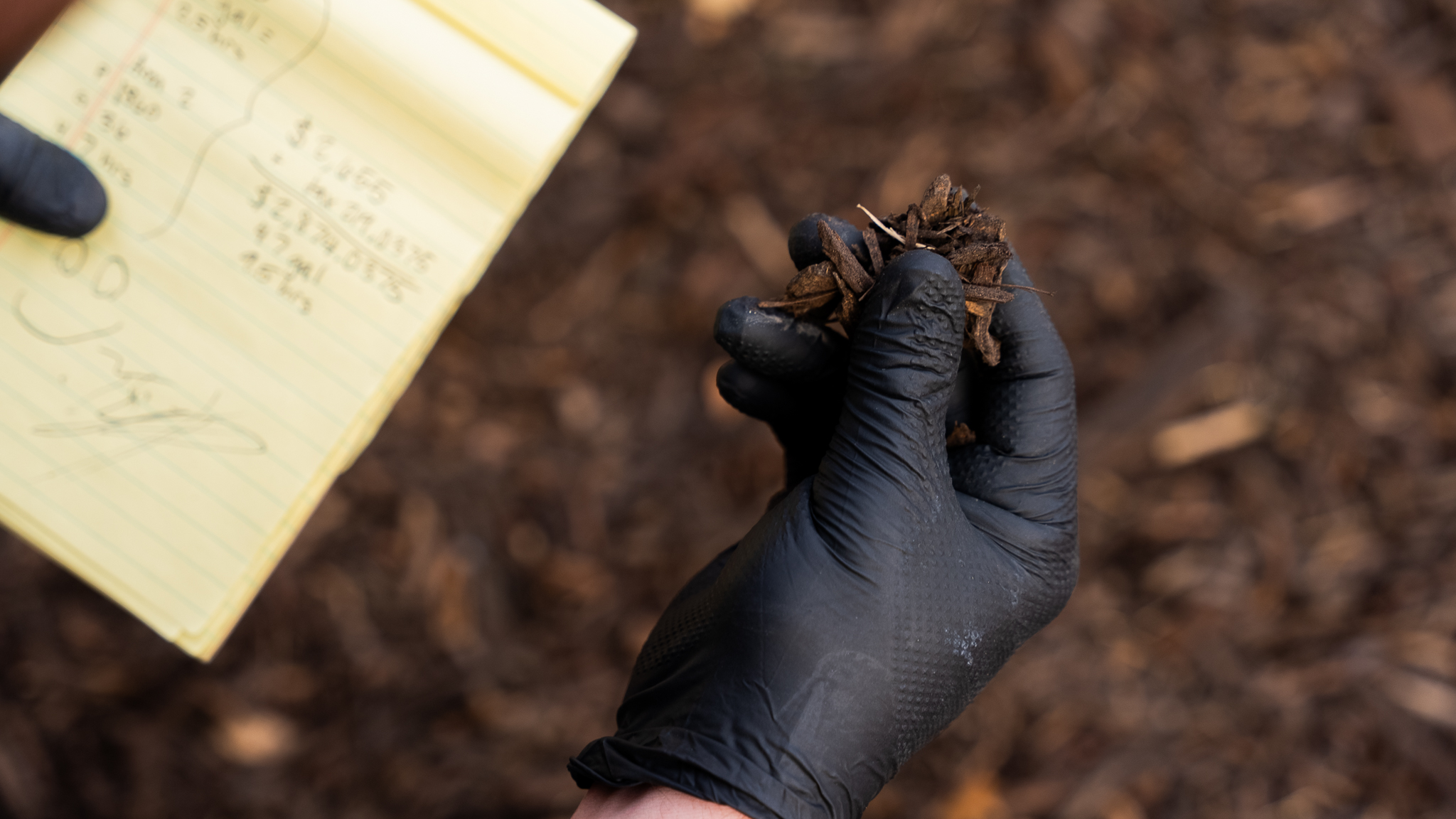 Gloved hand holds a small clump of dark soil and wood chips outdoors, with a yellow notebook in the background.