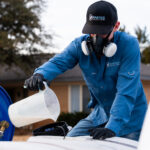 Worker in blue uniform and respirator fills a white container near a blue reel with red hose outdoors.