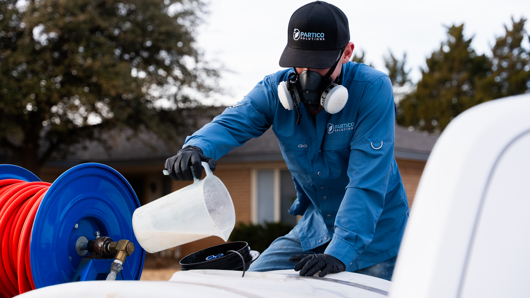 Worker in blue uniform and respirator fills a white container near a blue reel with red hose outdoors.