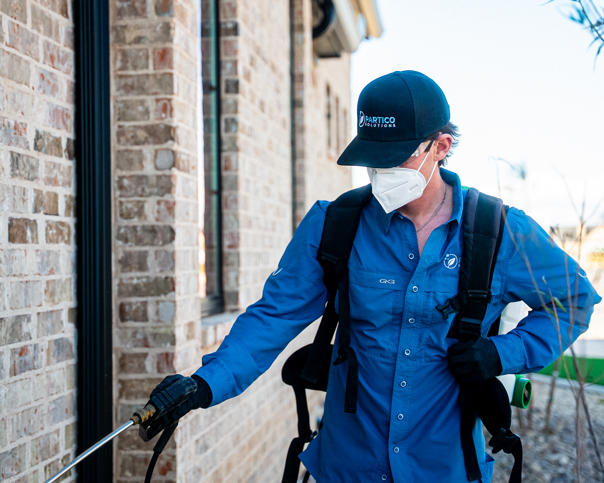 Worker in blue uniform wearing a white face mask operates a spray wand from a backpack sprayer beside a brick wall outside.