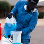 Person in a blue shirt and safety mask pours bright blue liquid from a gallon jug into a measuring cup on the back of a white vehicle.