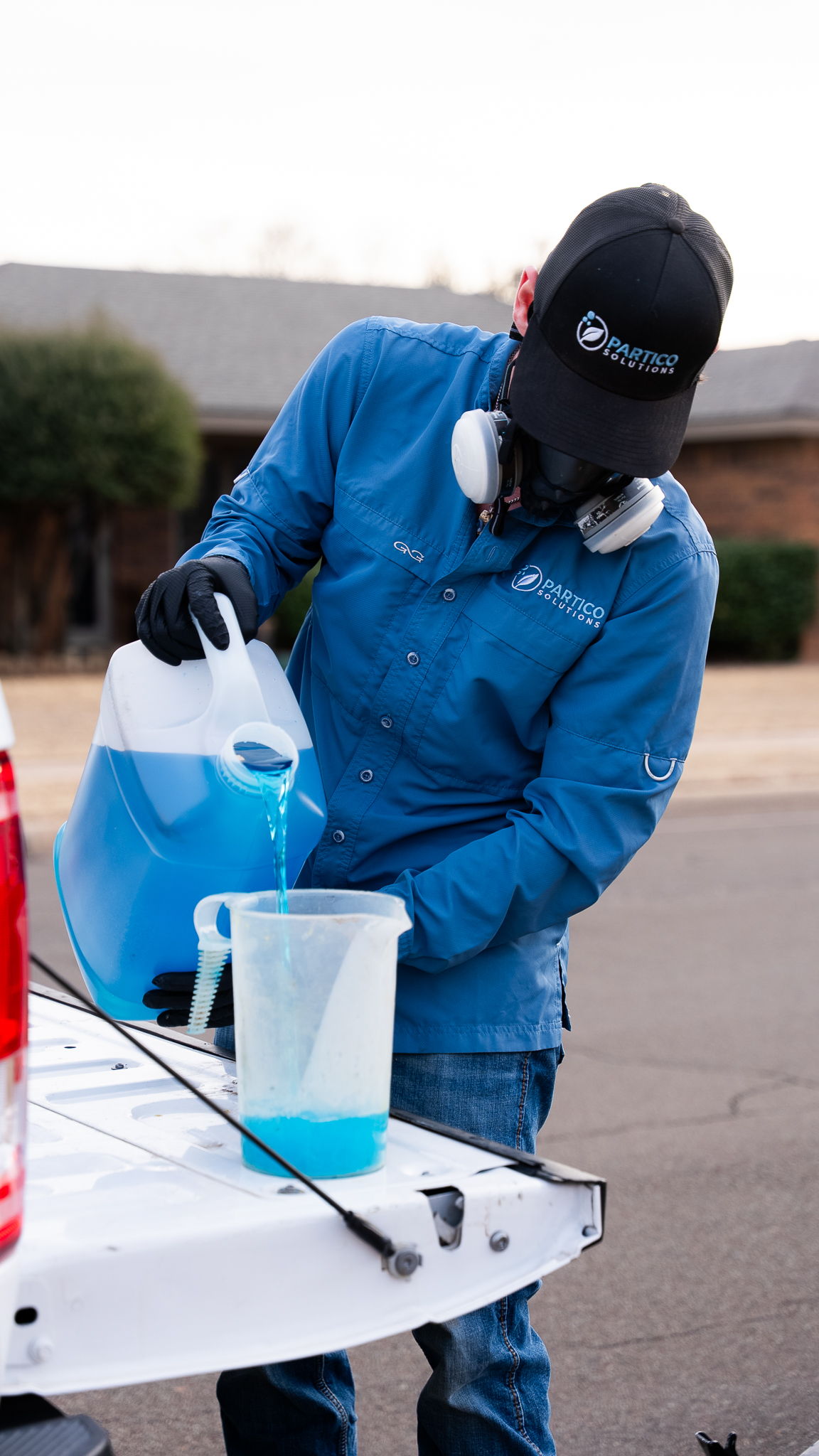 Person in a blue shirt and safety mask pours bright blue liquid from a gallon jug into a measuring cup on the back of a white vehicle.