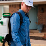 Pest control technician in a blue shirt and white cap with a backpack sprayer, standing outside a house, holding a spraying wand.