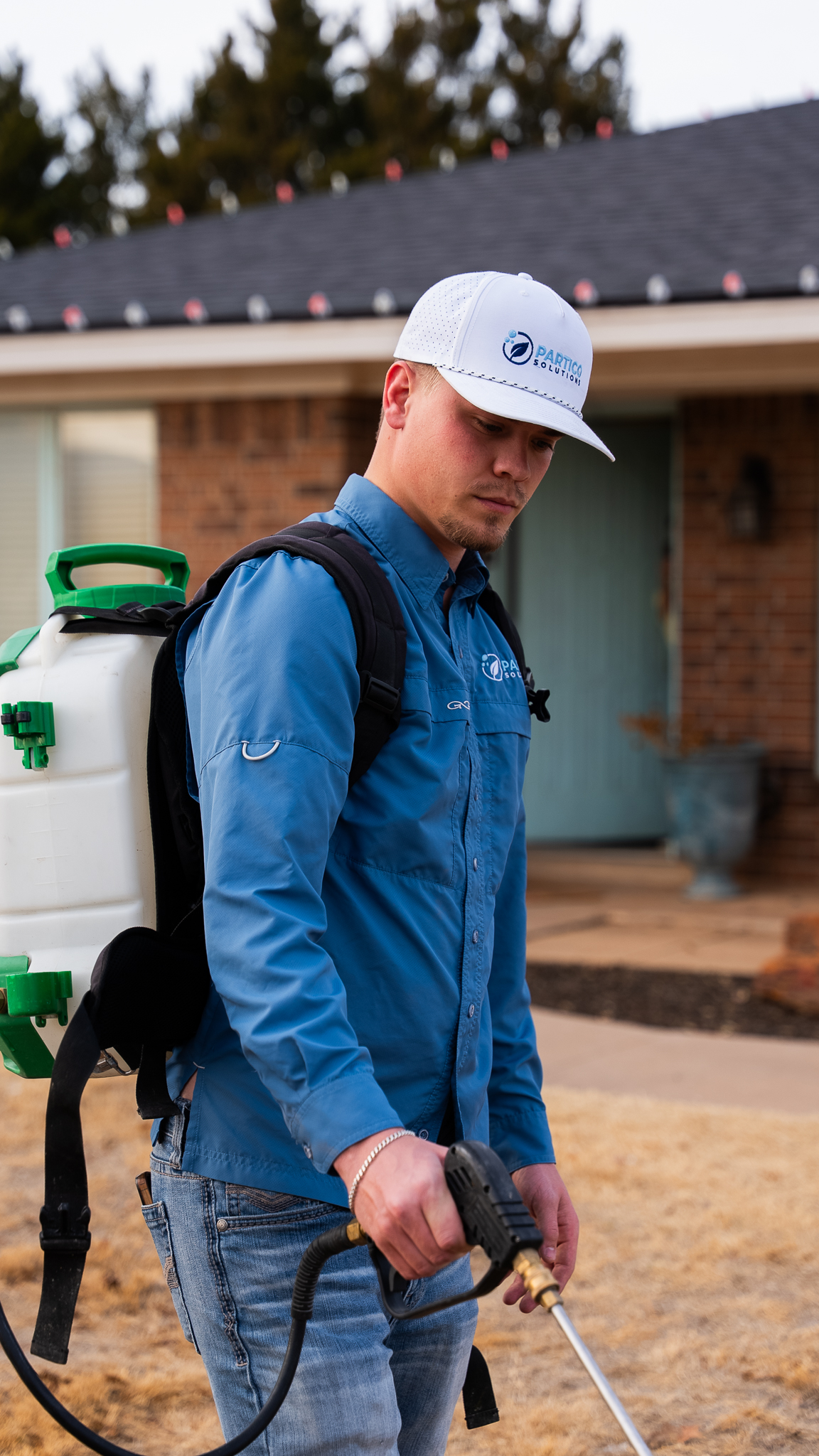 Pest control technician in a blue shirt and white cap with a backpack sprayer, standing outside a house, holding a spraying wand.