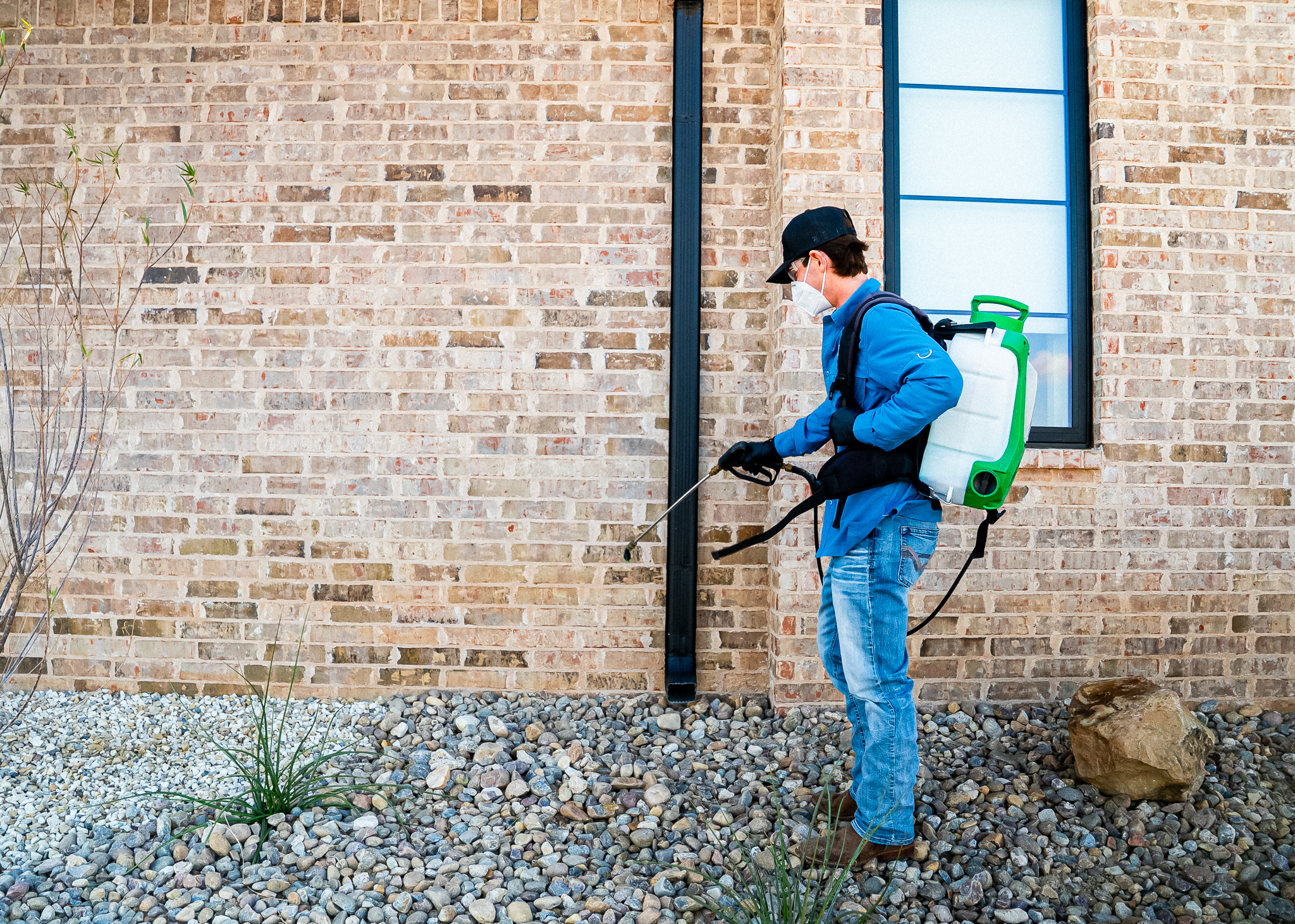 Pest control worker wearing a mask and blue uniform with a backpack sprayer spraying near a brick wall outdoors