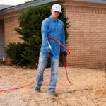 Worker in a blue shirt and white cap spraying with a red hose in a dry yard beside a brick house, wearing safety glasses.