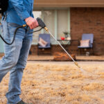 Person wearing a blue shirt and jeans uses a pressure washer to spray a driveway in front of a brick house.