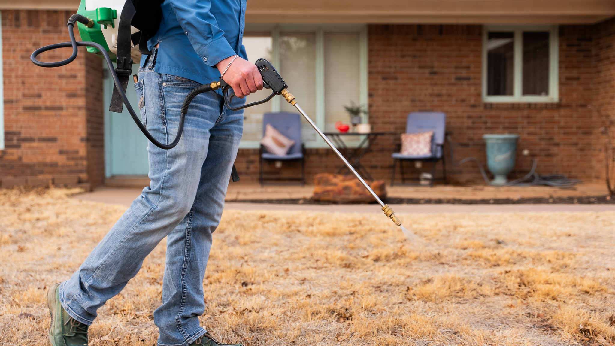 Person wearing a blue shirt and jeans uses a pressure washer to spray a driveway in front of a brick house.