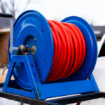 Blue hose reel with a neatly coiled bright red hose on a metal stand outdoors, close-up view focusing on the reel and crank handle.