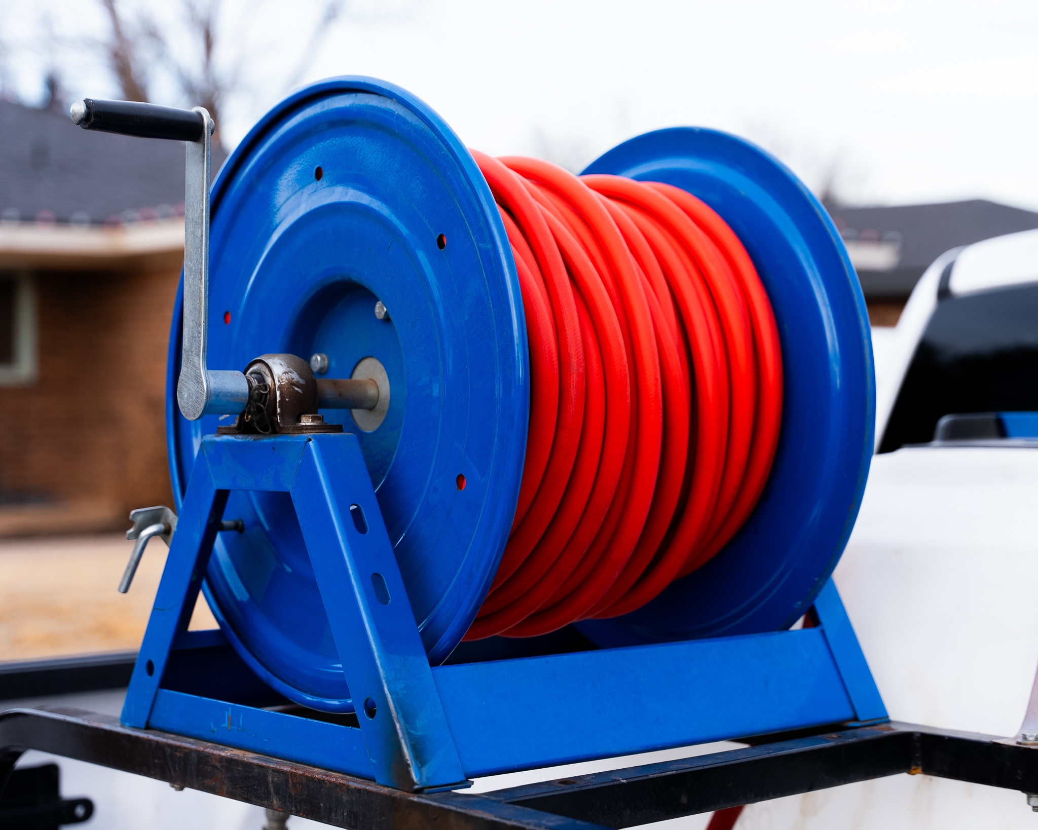Blue hose reel with a neatly coiled bright red hose on a metal stand outdoors, close-up view focusing on the reel and crank handle.