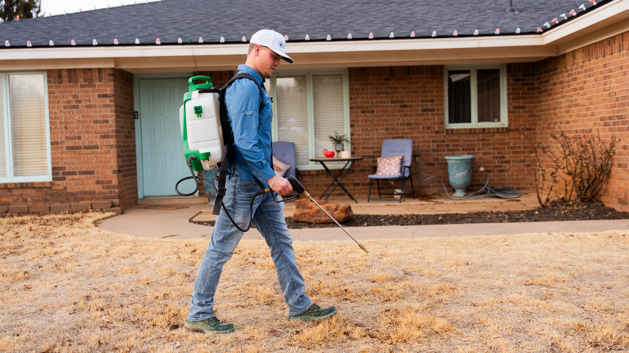 Maintenance worker with a white cap and blue shirt uses a green-and-white backpack sprayer to treat the front yard of a brick house in a suburban setting.