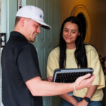 Man wearing a white cap hands a tablet to a smiling woman at the doorway of a brick house.