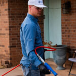 Man in a blue work shirt and white cap uses a blue pressure washer outside a brick house, spraying water.