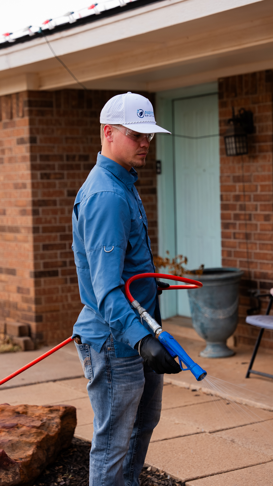 Man in a blue work shirt and white cap uses a blue pressure washer outside a brick house, spraying water.