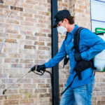 Worker in blue uniform, safety mask and goggles, operating a pressure washer on a brick wall beside a glass door.