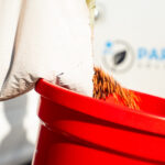 Close-up of a white package with green text being dropped into a bright red trash bin, with shredded packing material spilling over the rim.