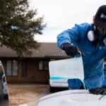 Worker in blue coveralls and respirator applies a coating to a car hood in a driveway.