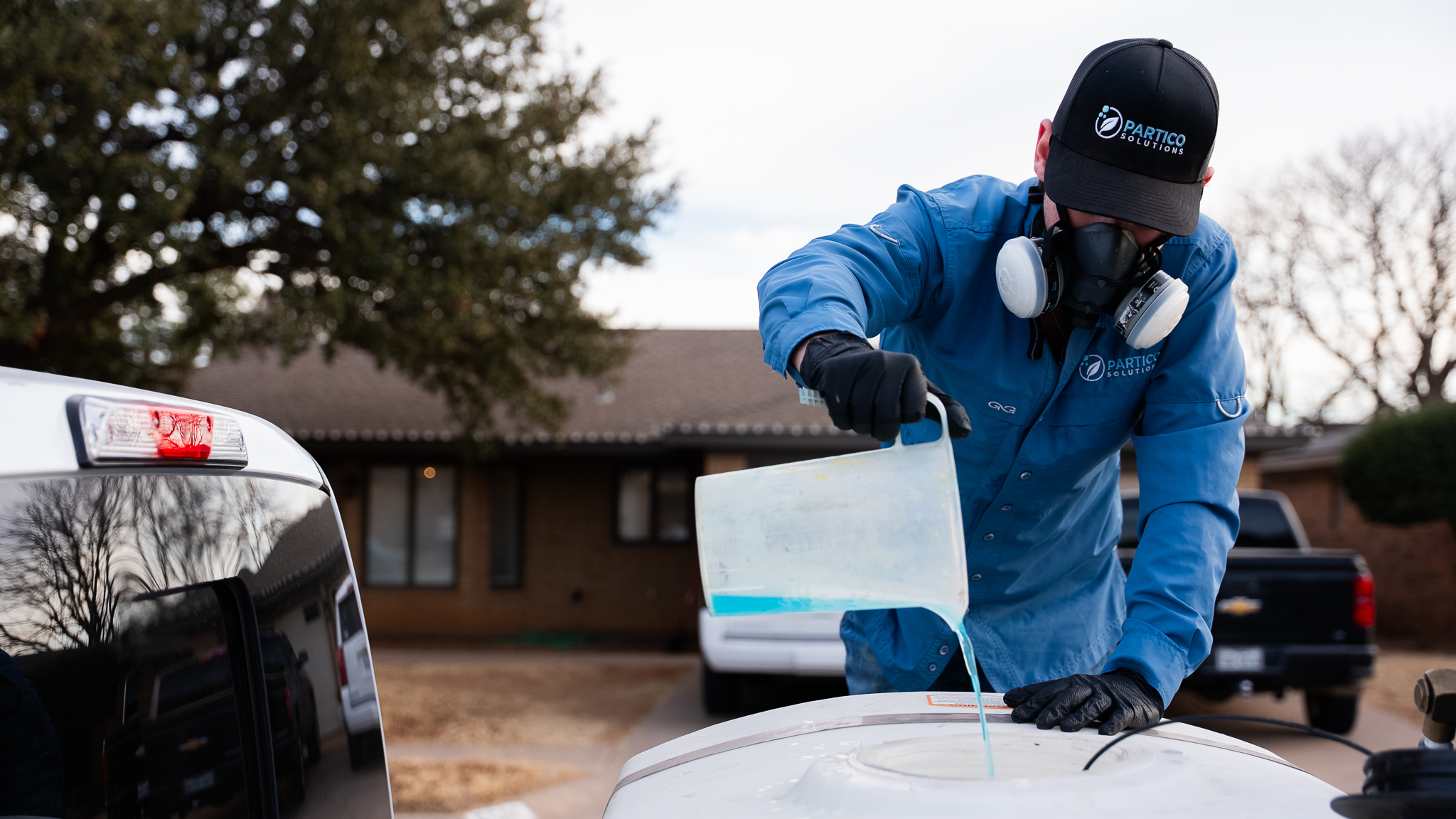 Worker in blue coveralls and respirator applies a coating to a car hood in a driveway.