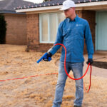 Man in a blue work shirt and white cap holds a red hose with a spray nozzle in a dry front yard by a brick house.