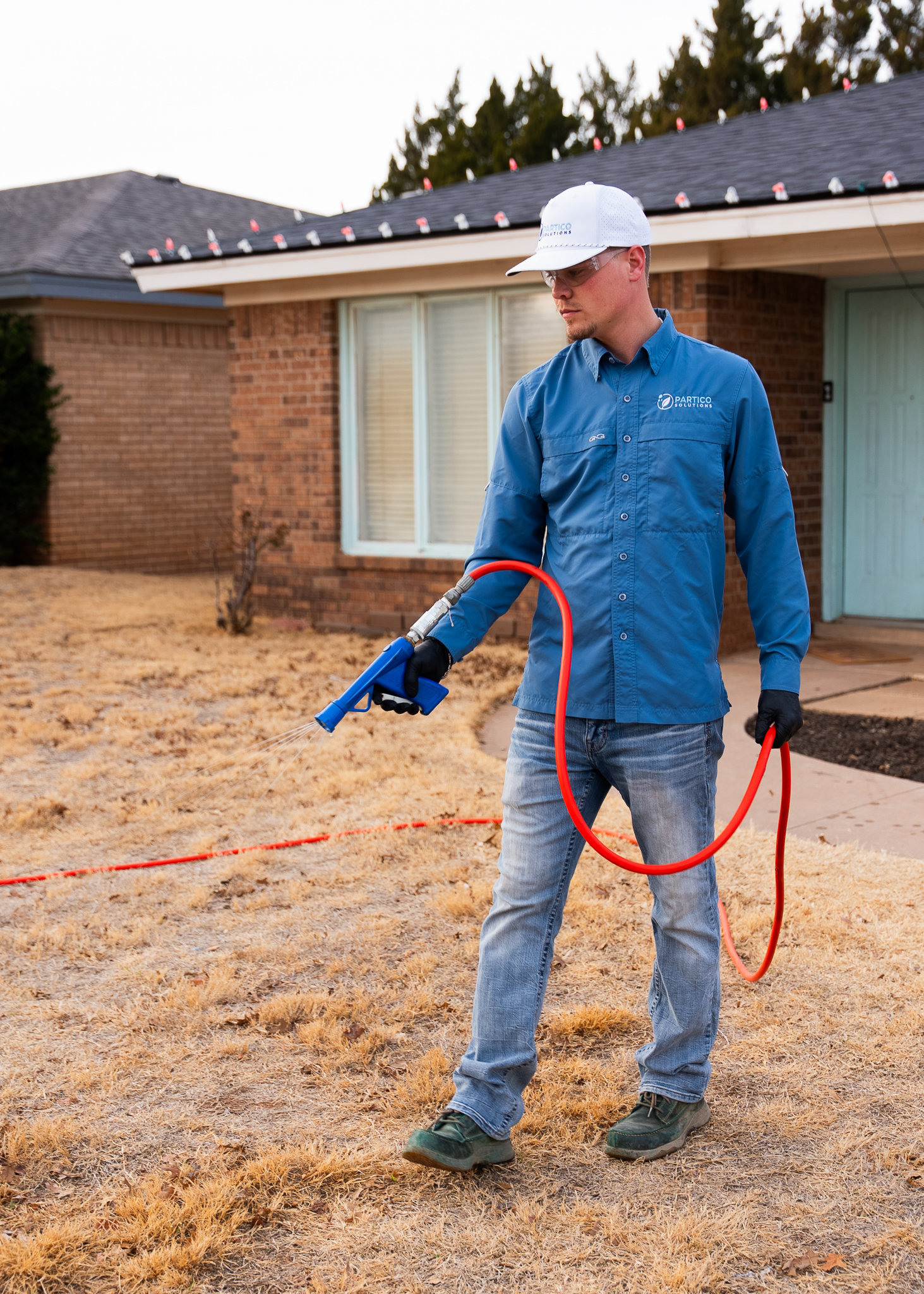 Man in a blue work shirt and white cap holds a red hose with a spray nozzle in a dry front yard by a brick house.