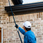 Maintenance worker in a blue shirt and white cap uses an extending pole with a yellow cleaning head to clean a brick wall outside.