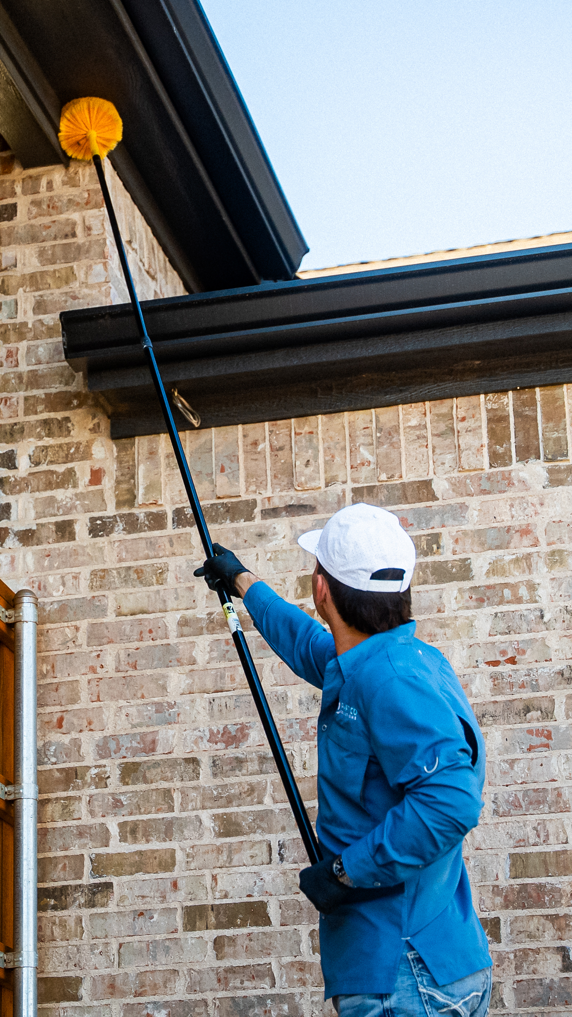 Maintenance worker in a blue shirt and white cap uses an extending pole with a yellow cleaning head to clean a brick wall outside.