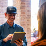 Technician in a blue uniform and cap using a tablet indoors near brick columns, smiling at the screen.