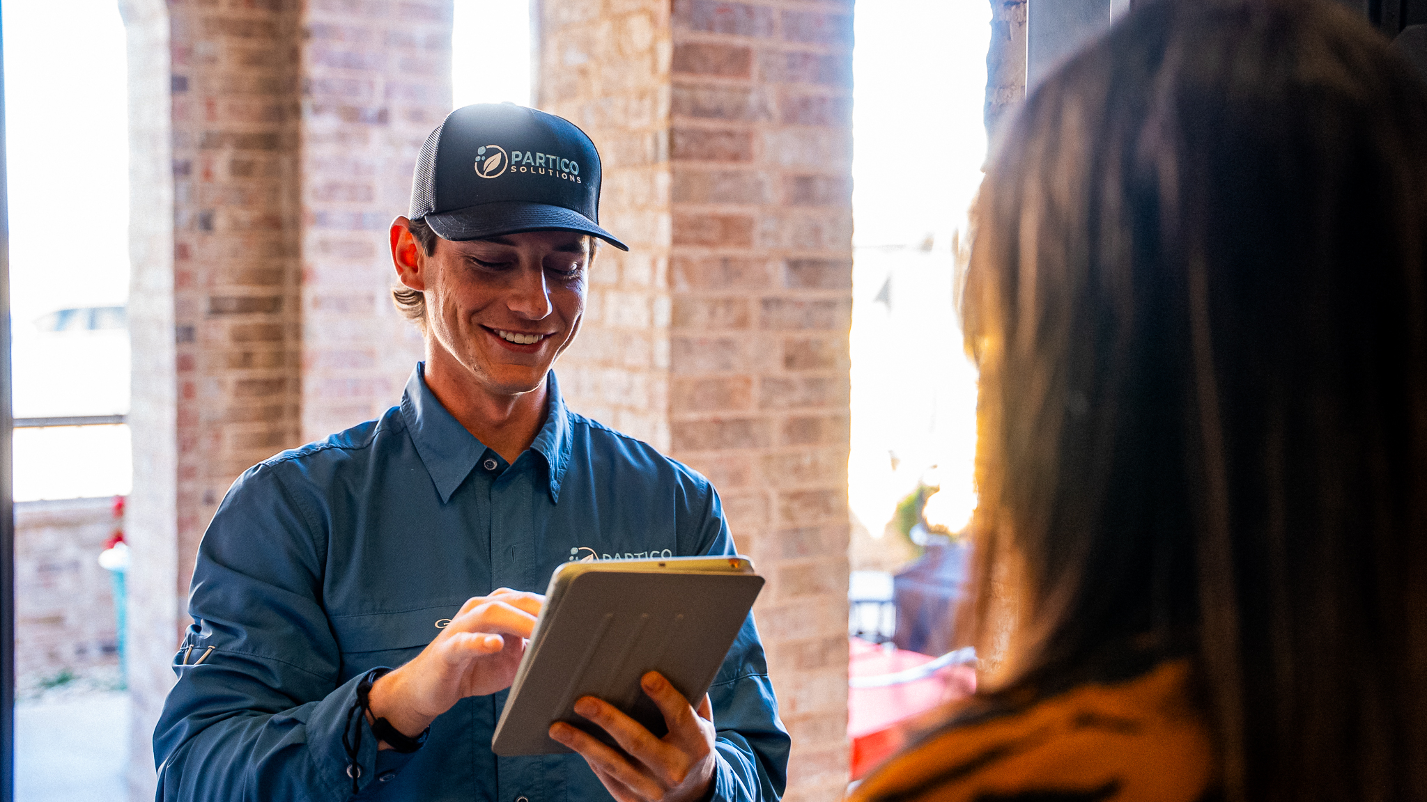 Technician in a blue uniform and cap using a tablet indoors near brick columns, smiling at the screen.