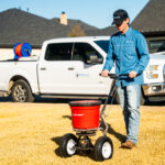Man in a blue shirt and cap pushes a small red EarthWay seed spreader on a sunny lawn with a white pickup truck in the background.