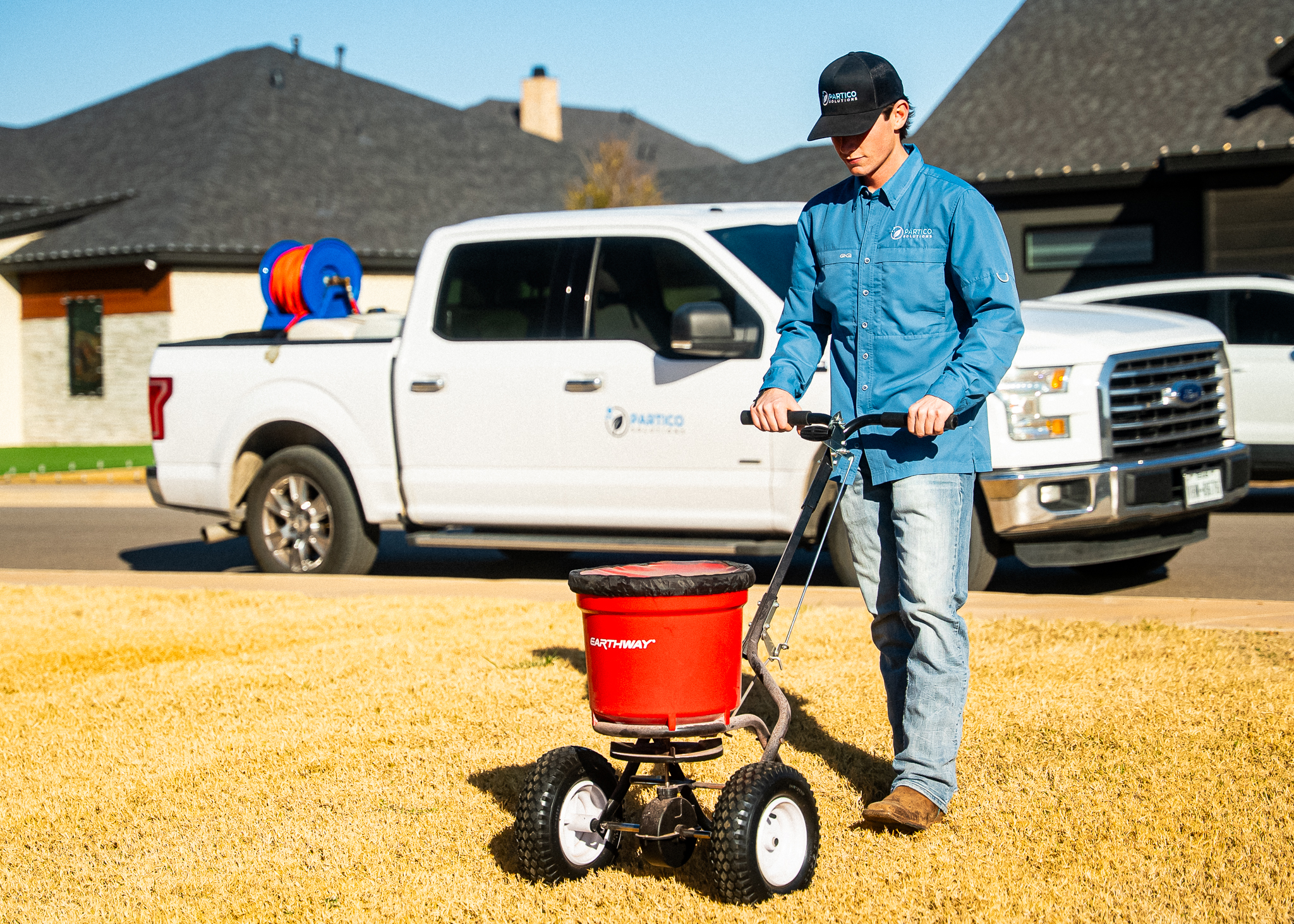 Man in a blue shirt and cap pushes a small red EarthWay seed spreader on a sunny lawn with a white pickup truck in the background.