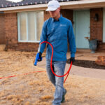 Man in a blue work shirt and white cap holding a red hose nozzle outdoors in front of a brick house.