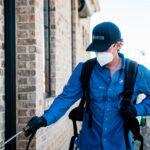 Worker in blue uniform, mask, and helmet uses a spray wand to clean or treat a brick exterior wall.
