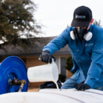 Worker in a blue uniform and respirator mask handles a white container near a blue hose reel outdoors, with a building in the background