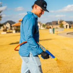 Man in a blue shirt and light jeans sweeping the ground with a blue–yellow broom outdoors in a sunny, suburban area.