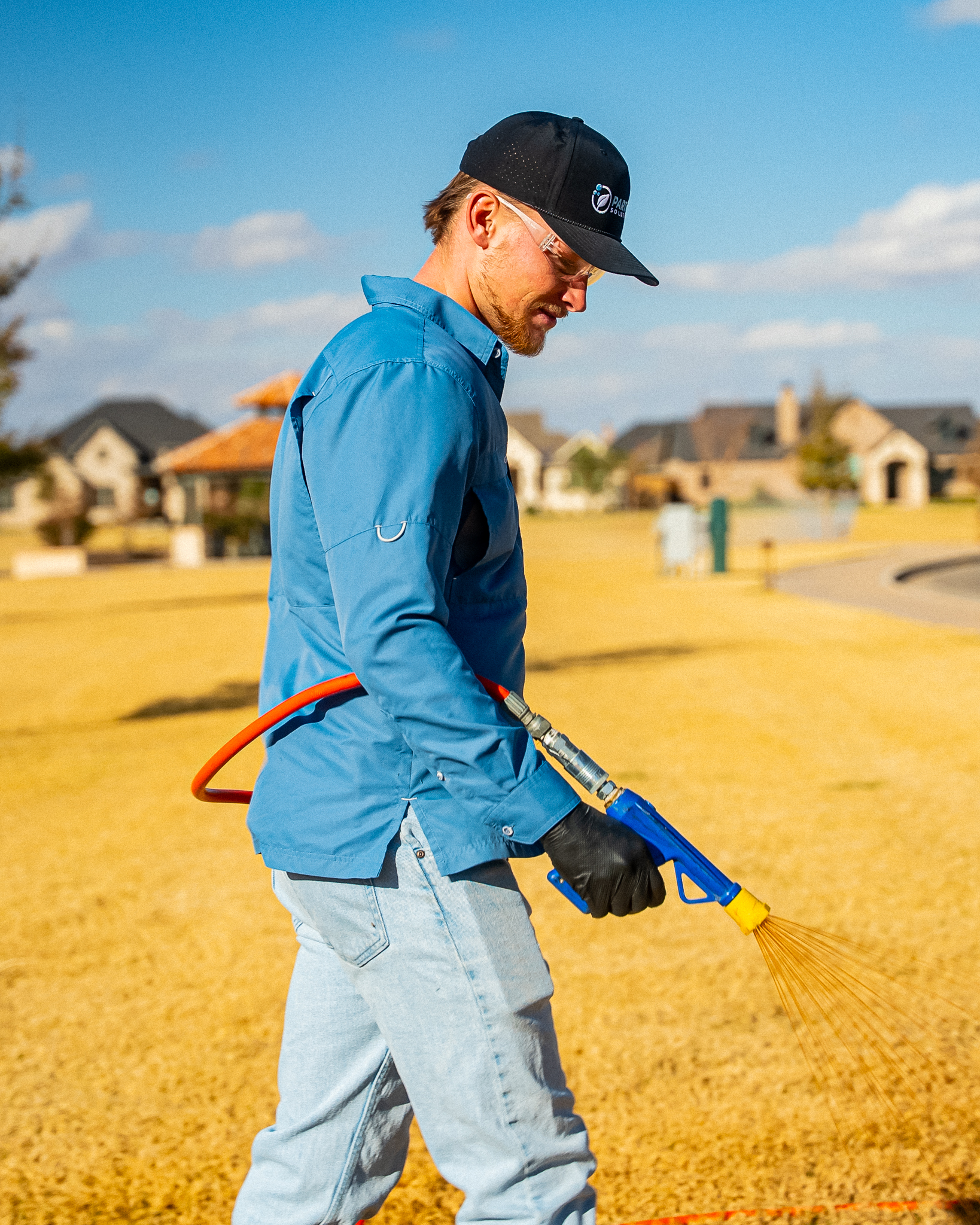 Man in a blue shirt and light jeans sweeping the ground with a blue–yellow broom outdoors in a sunny, suburban area.