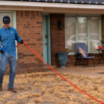 Man in a blue uniform and cap stands on a front lawn, holding an orange extension cord with a blue tool attached.