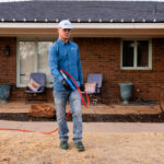 Man in a blue work shirt and white cap holding a red hose in front of a brick house, appearing ready to power wash the porch area.