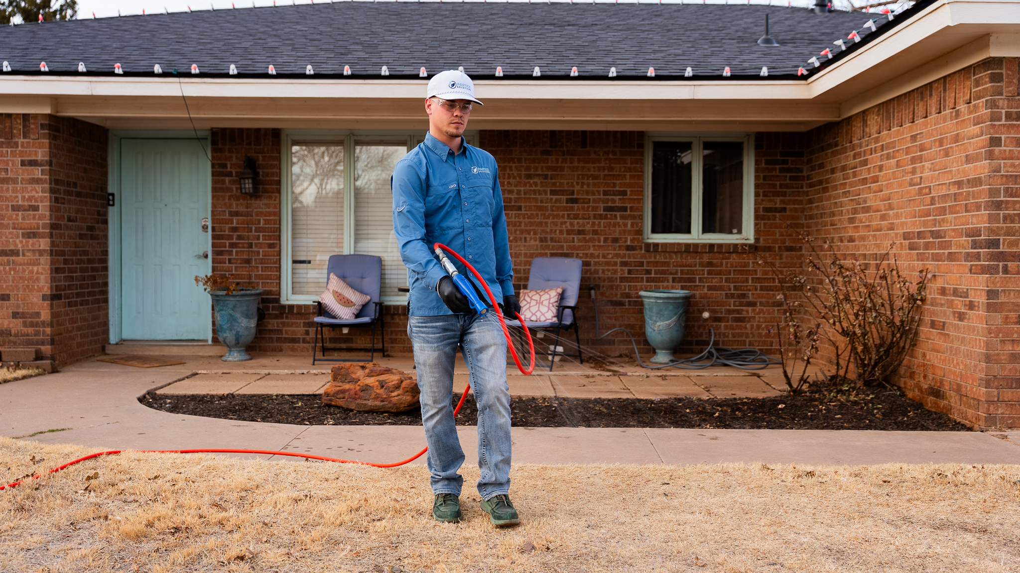 Man in a blue work shirt and white cap holding a red hose in front of a brick house, appearing ready to power wash the porch area.